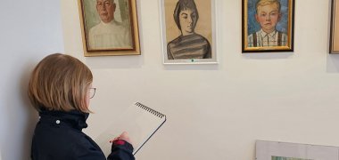 Wand mit Bilder; davor steht eine Frau und Zeichnet auf einem Block