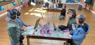 Kinder stehen vor einem Tisch und hören Musik über Kopfhörer