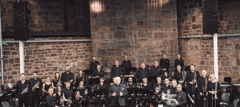 Gruppenaufnahme der Brass Band Hessen auf einer Bühne vor einer historischen Natursteinmauer. Die Musikerinnen und Musiker sind in mehreren Reihen mit ihren Blechblasinstrumenten und Schlagwerk aufgestellt. Im Vordergrund steht Dirigent Hans-Reiner Schmidt. Alle tragen überwiegend schwarze Konzertkleidung.