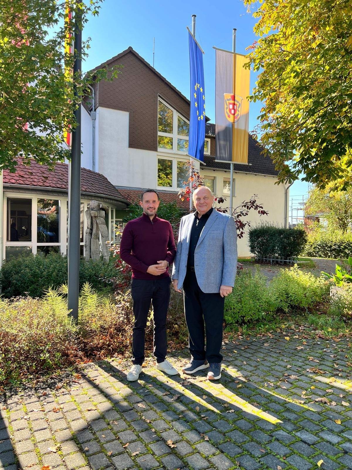 Bürgermeister Luca Fritsch (links) und Dirigent Hans-Reiner Schmidt (rechts) stehen nebeneinander auf dem gepflasterten Vorplatz des Rathauses Willingshausen. Im Hintergrund sind das Rathaus, eine Europaflagge und die Gemeindeflagge zu sehen.
